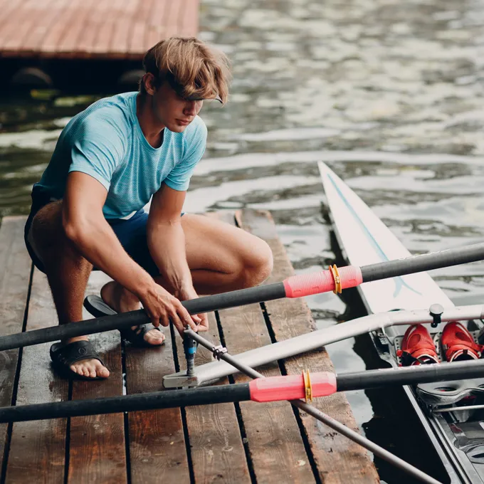 Un jeune homme en t-shirt de sport bleu répare des rames sur un quai au bord de l'eau.
