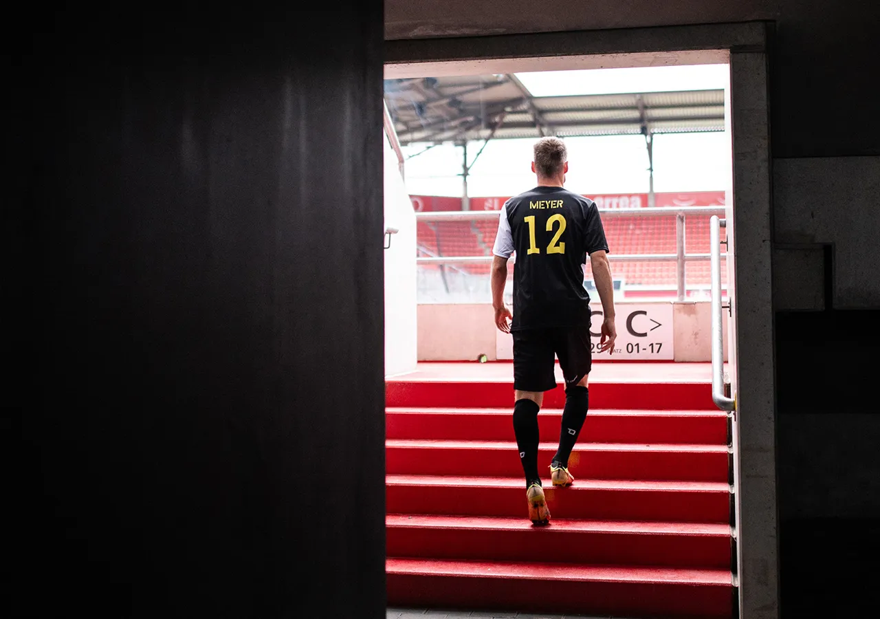Un joueur de football portant un maillot avec le nom Meyer et le numéro 12 monte un escalier rouge vers un stade.