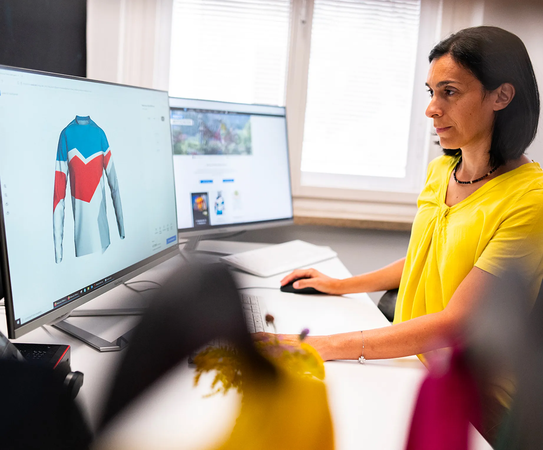 Une femme en haut jaune est assise à un bureau et conçoit sur un écran d'ordinateur un maillot de sport personnalisé avec des panneaux bleus, rouges et blancs.
