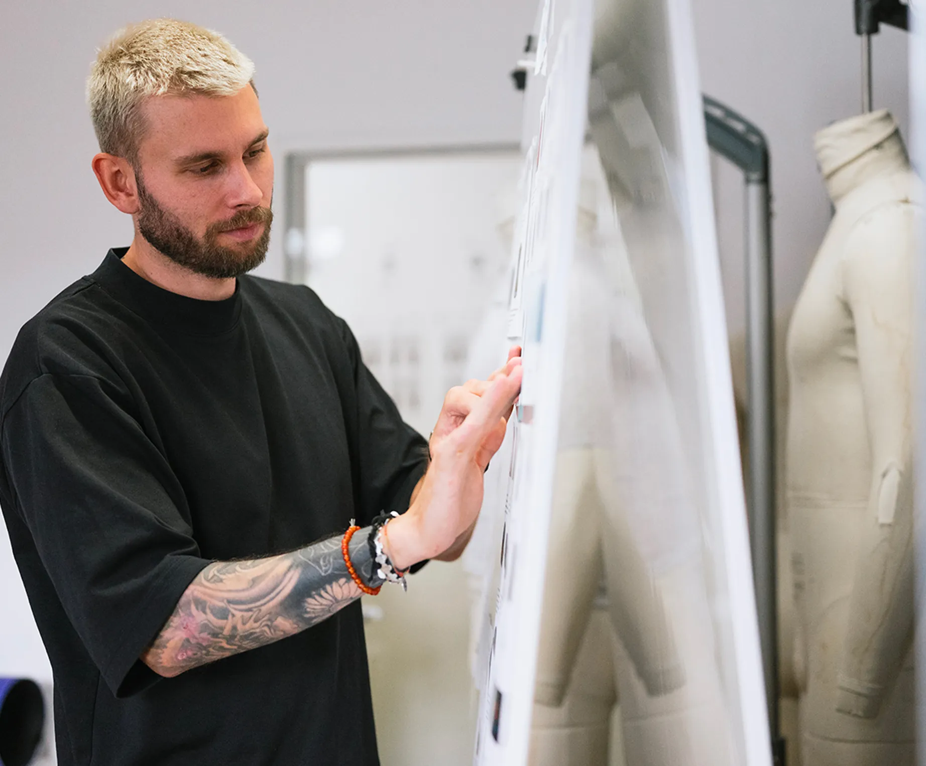 Un homme aux cheveux blonds et avec un tatouage au bras examine attentivement des dessins sur un tableau d'affichage dans un studio de design, une poupée de couture étant visible en arrière-plan.